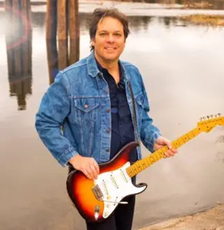 A man holding a guitar in front of water.