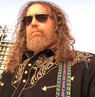 A man with long hair and sunglasses is wearing a black shirt.