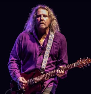 A man with long hair and a purple shirt is playing an electric guitar.