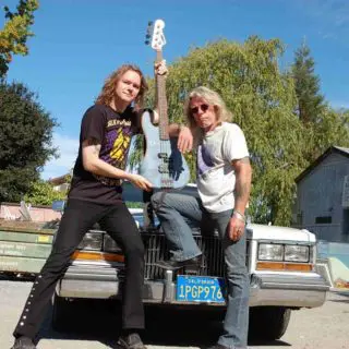 Two men are posing on a car with a guitar.